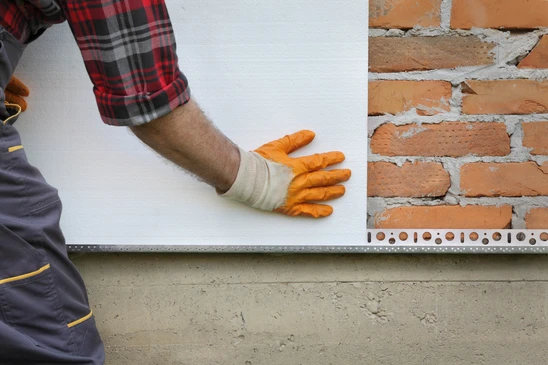 Ouvrier en combinaison et gants orange appliquant isolant polystyrène blanc sur mur extérieur en briques apparentes